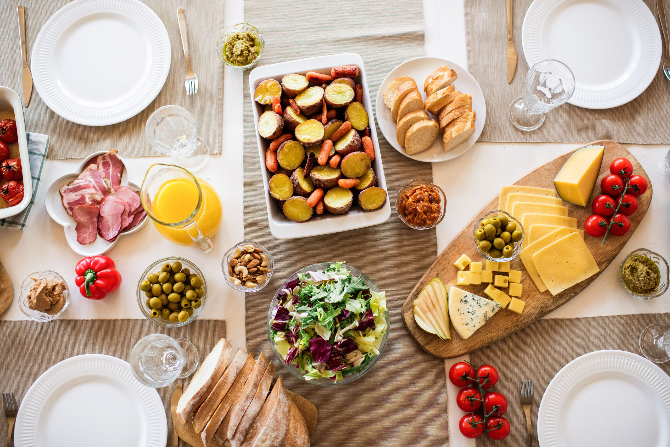 A breakfast table seen from above with cheese, ham, bread, roasted vegetables, salad and juice