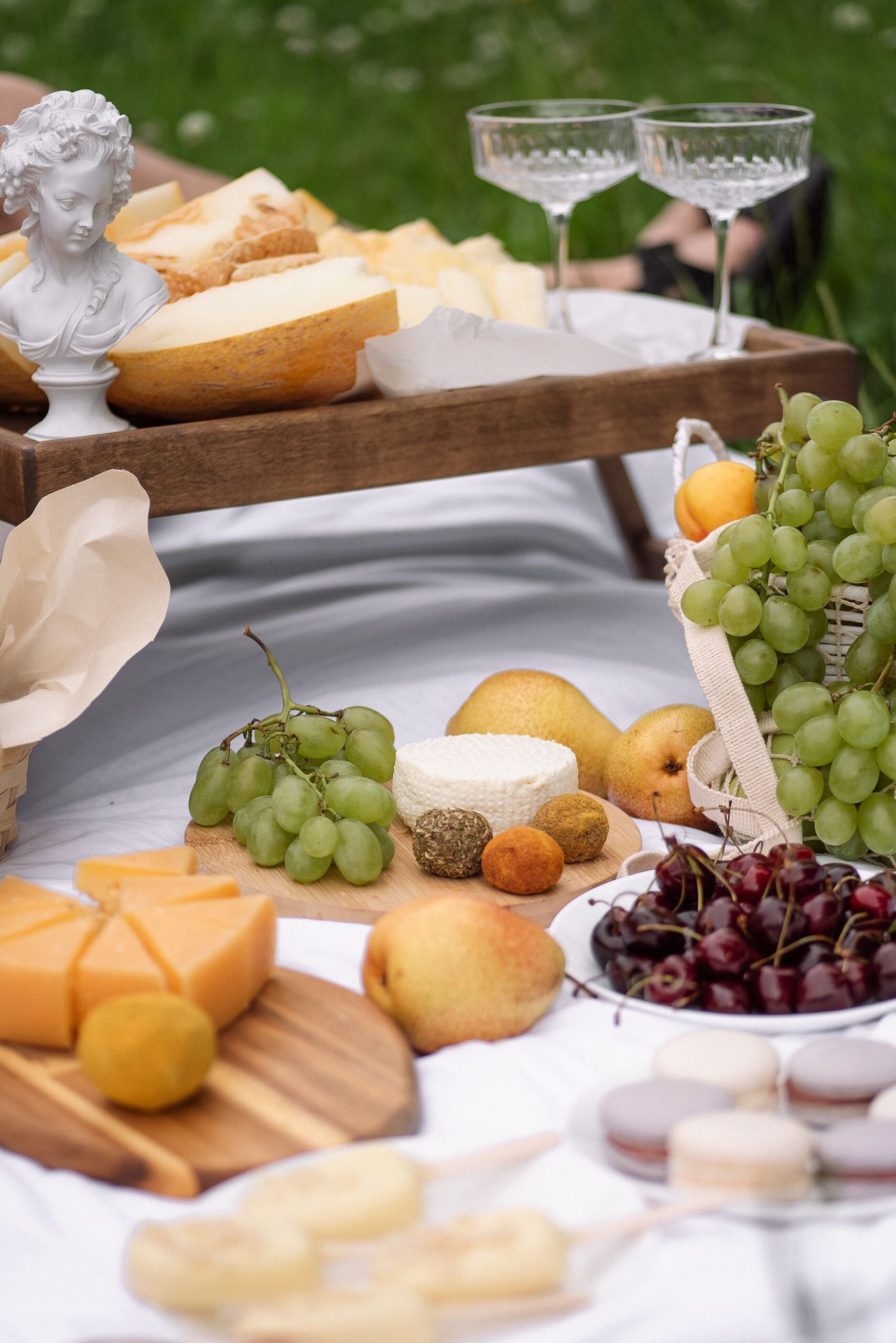 A picnic of cheese, fruit, bread and glasses on a white cloth in the grass