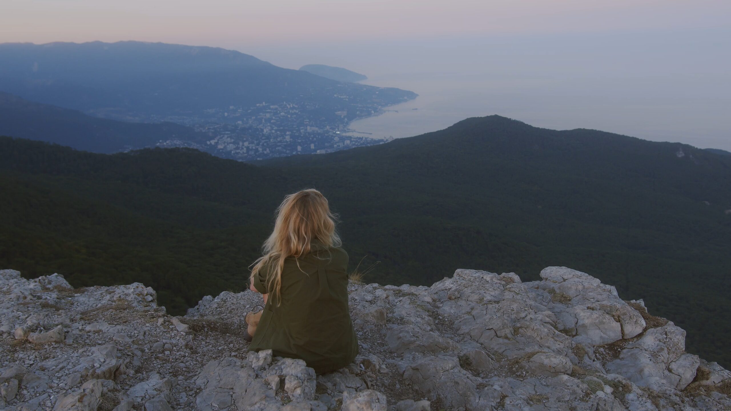 A woman sits on a rock above a wooded valley at dusk